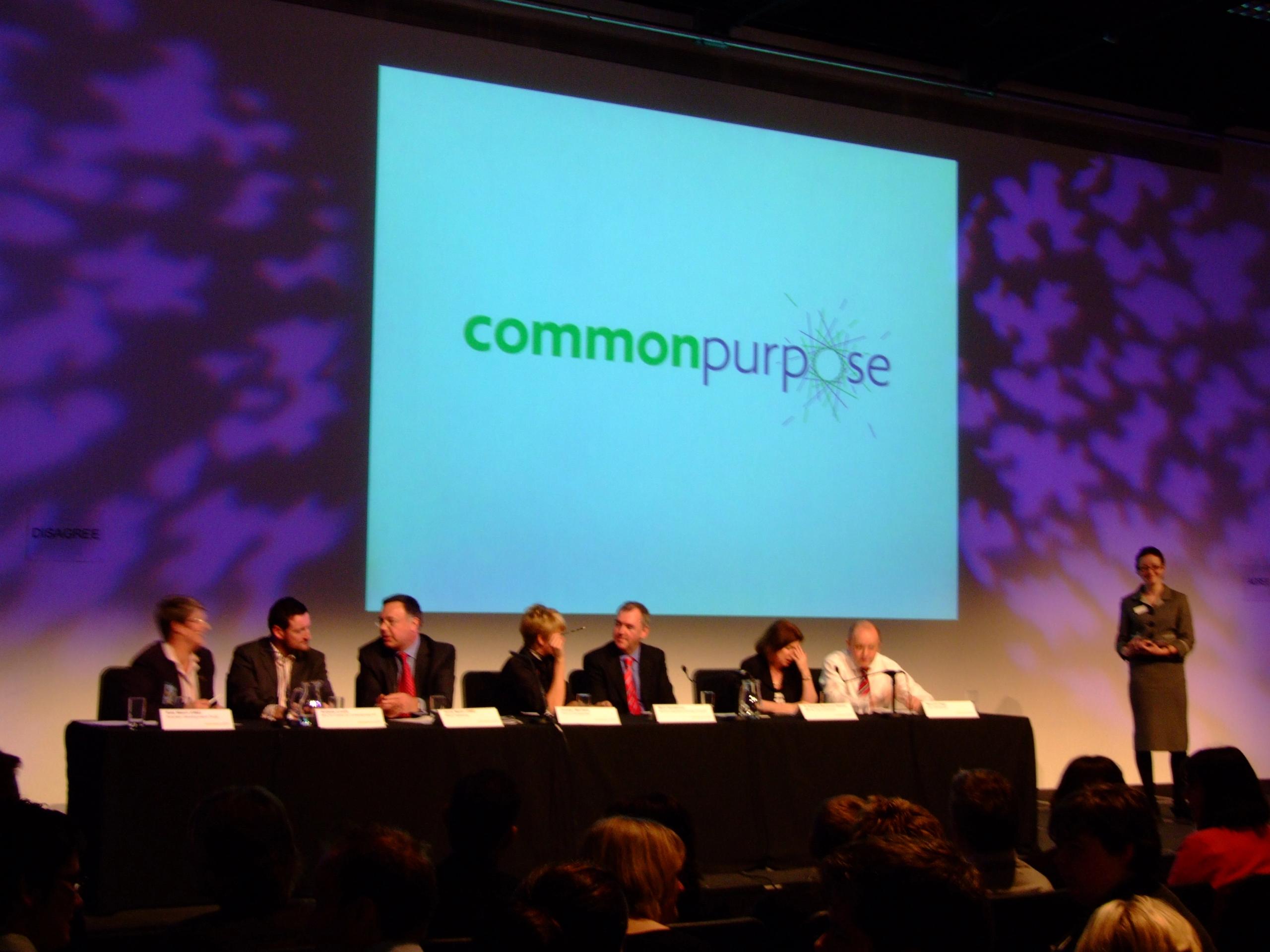 A row of people sitting on a stage beneath a projector screen with the Common Purpose logo